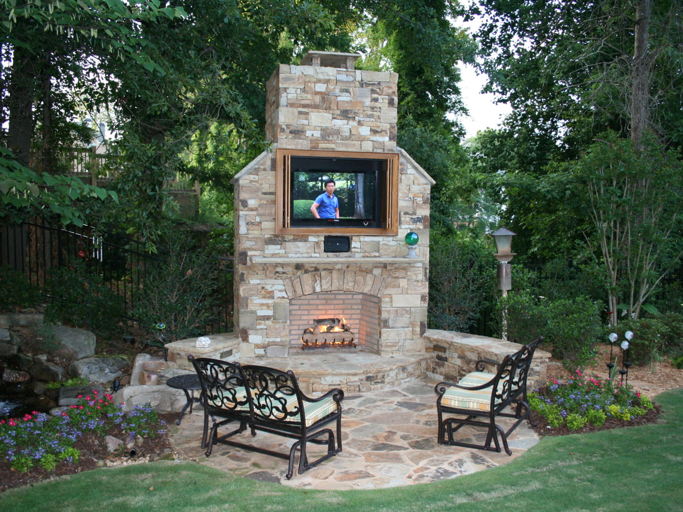 An inviting outdoor patio features a stone fireplace with a TV mounted above it. Nearby, an outdoor kitchen adds to the allure, while two benches face the warmth of the fire, surrounded by lush trees and garden landscaping.