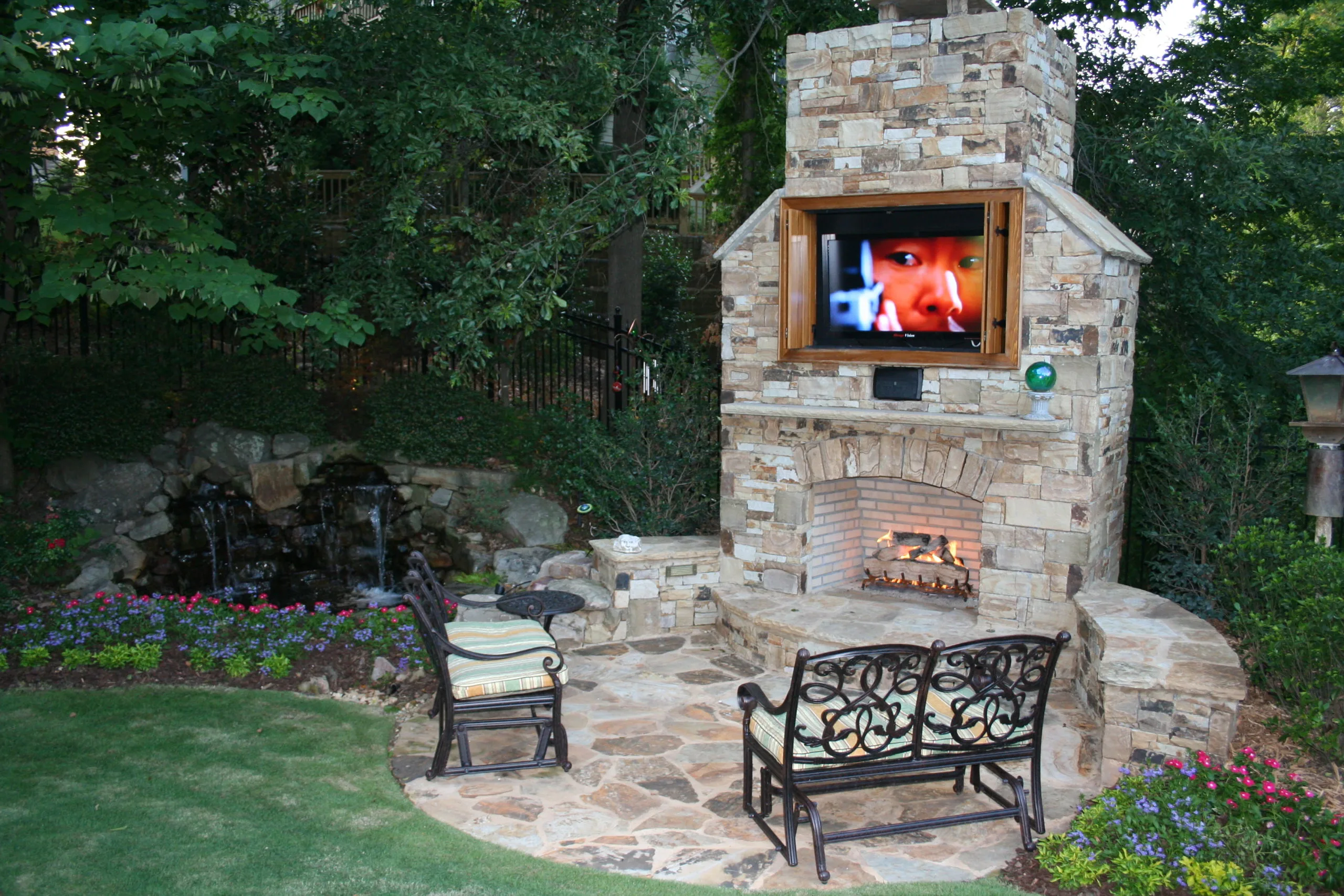 An outdoor patio with a stunning stone fireplace, complete with a TV above, is complemented by elegant wrought iron furniture. A tranquil waterfall feature surrounded by lush greenery and vibrant flowers creates a serene escape. An outdoor patio with a stunning stone fireplace, complete with a TV above, is complemented by elegant wrought iron furniture. A tranquil waterfall feature surrounded by lush greenery and vibrant flowers creates a serene escape.