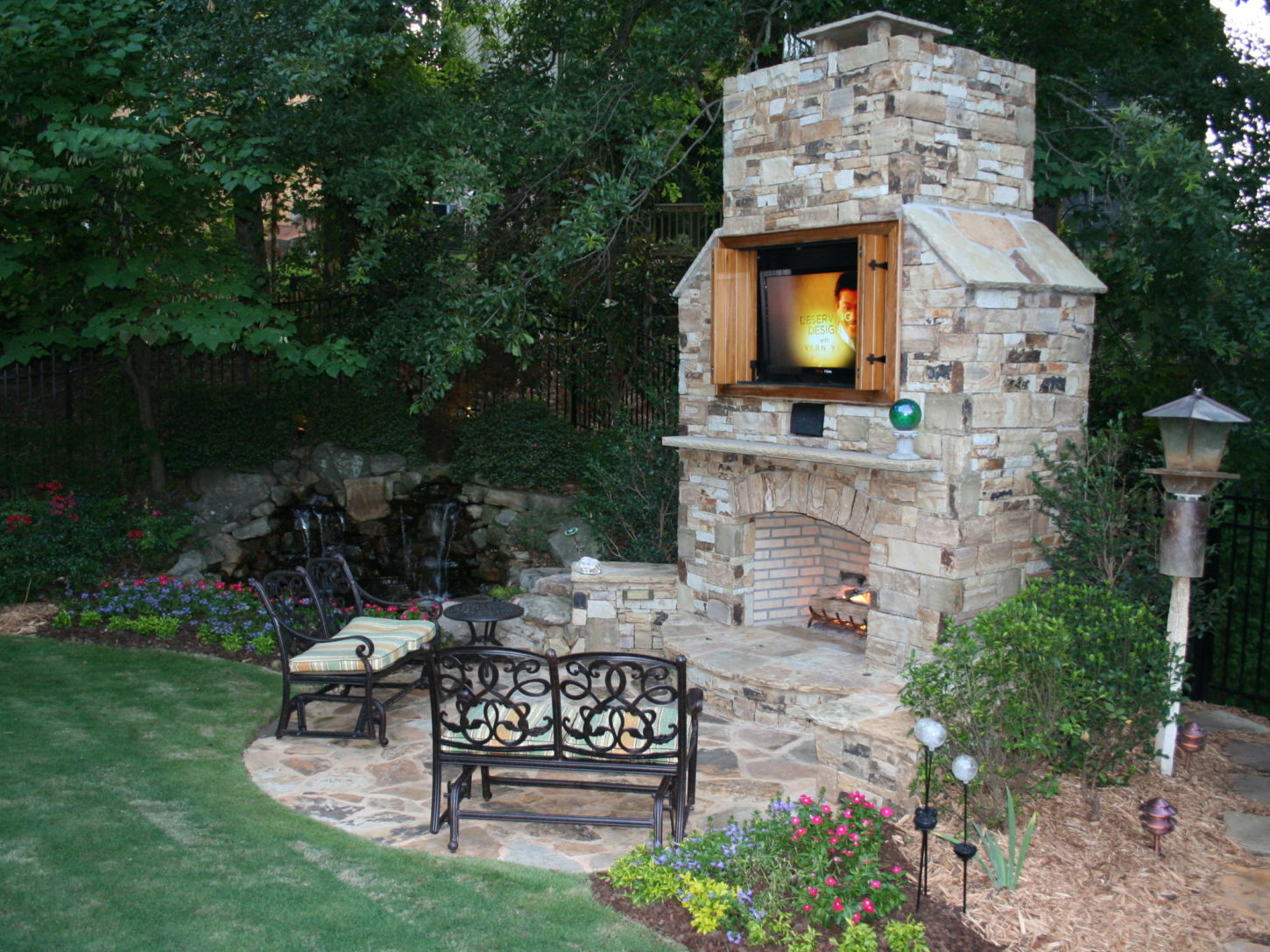 An outdoor stone fireplace with a built-in TV anchors this enchanting space, complemented by metal chairs and blooming flowers. The adjacent outdoor kitchen enhances the garden area, inviting you to unwind amidst lush greenery and culinary delights.
