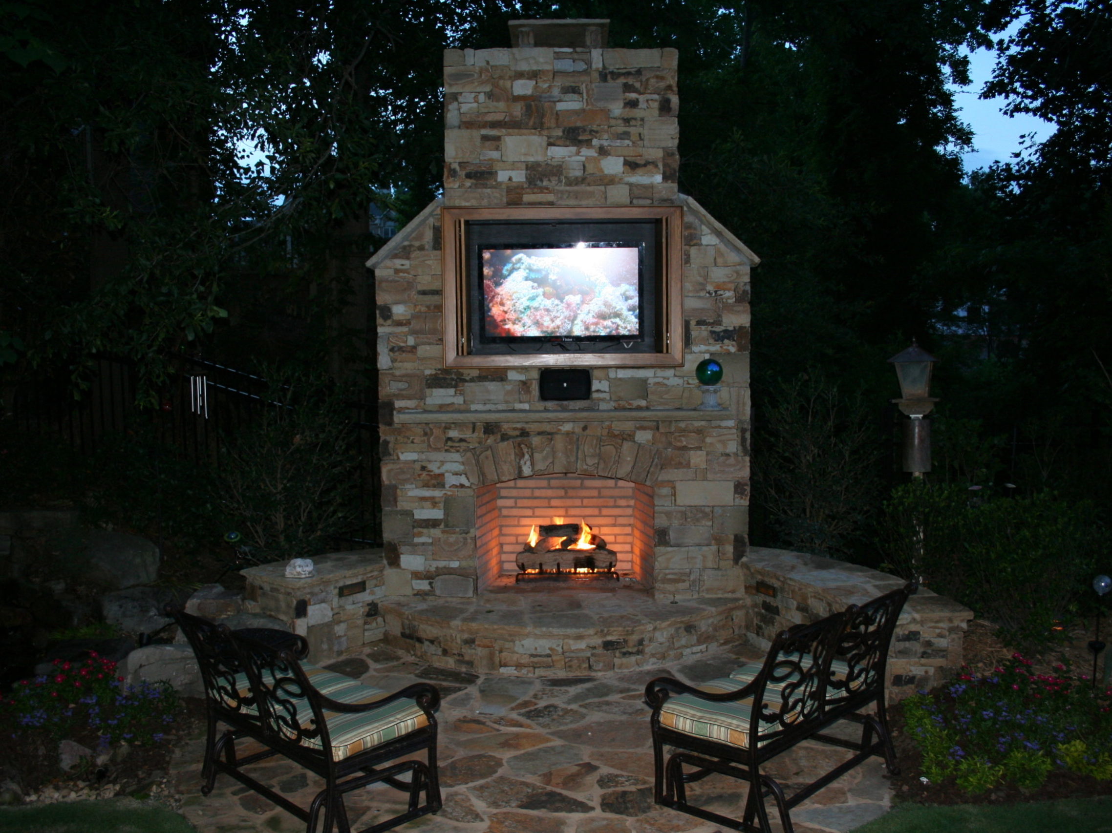 An outdoor stone fireplace with a TV mounted above is the centerpiece, complemented by an inviting outdoor kitchen. Flanked by two cushioned chairs, this setting boasts evening lighting and lush surrounding greenery, perfect for relaxation and entertainment.