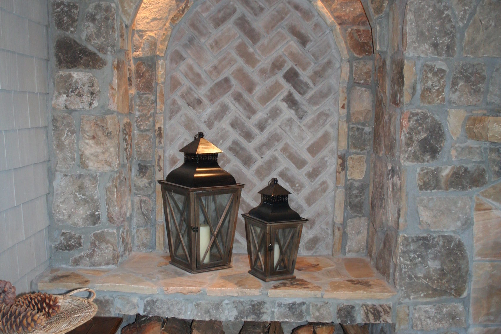 A charming stone alcove enhances your outdoor living space, featuring stacked firewood and two lanterns on a shelf. The herringbone brick pattern in the niche adds a touch of rustic elegance to this cozy retreat.