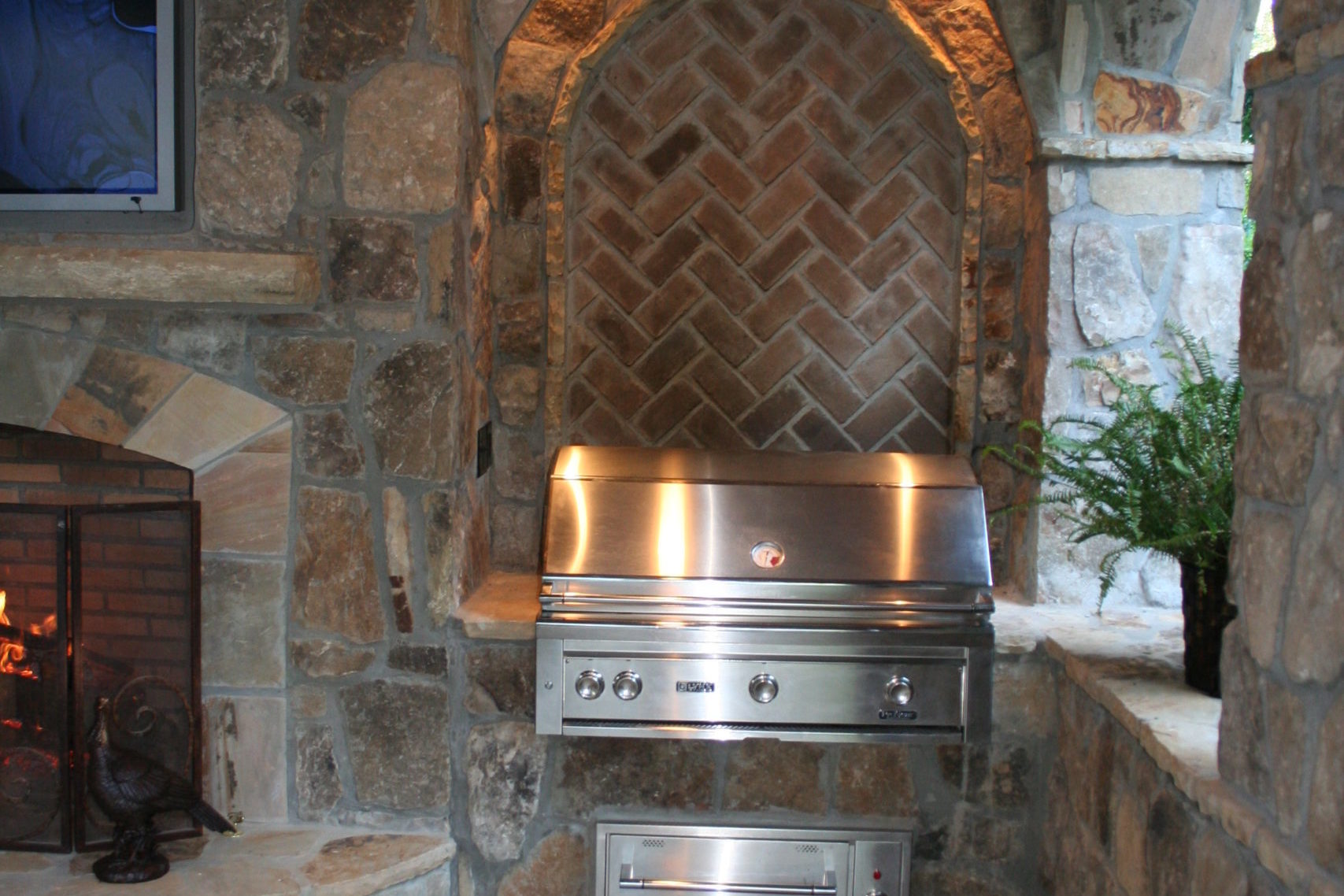Stone outdoor living kitchen area featuring a built-in stainless steel grill, arched brick backsplash, and a wood-fired oven to the left.