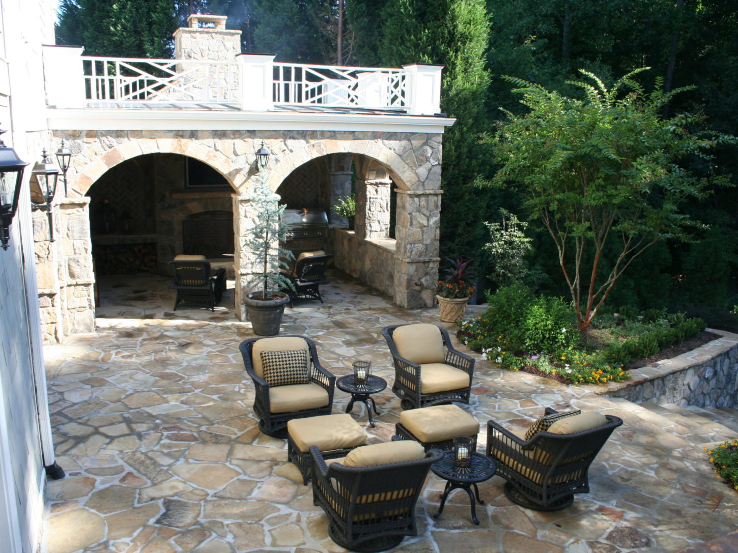 Experience the best in outdoor living with a spacious patio featuring stone flooring, wicker chairs with beige cushions, and a cozy fire pit. A stunning stone archway with a chimney completes this serene wooded setting.