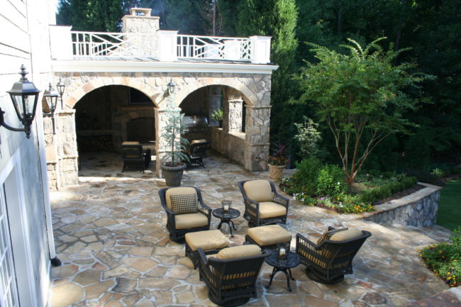 Experience the best in outdoor living with a spacious patio featuring stone flooring, wicker chairs with beige cushions, and a cozy fire pit. A stunning stone archway with a chimney completes this serene wooded setting.