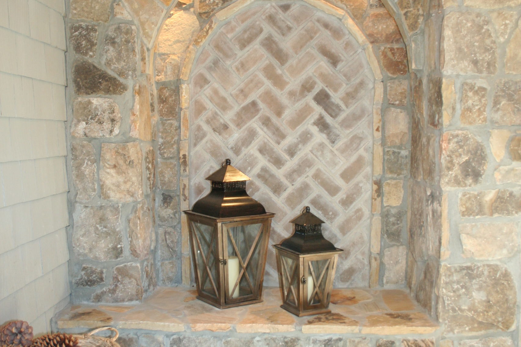 Experience outdoor living in this stone nook featuring a herringbone brick pattern. Two lanterns rest on a shelf above stacked firewood, casting a warm glow over the wooden table adorned with rustic pine cones on the side.