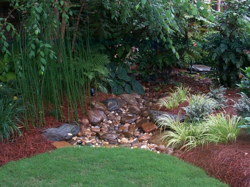 A serene garden featuring a small rock stream, surrounded by diverse plants, lush grasses, and a bed of mulch.