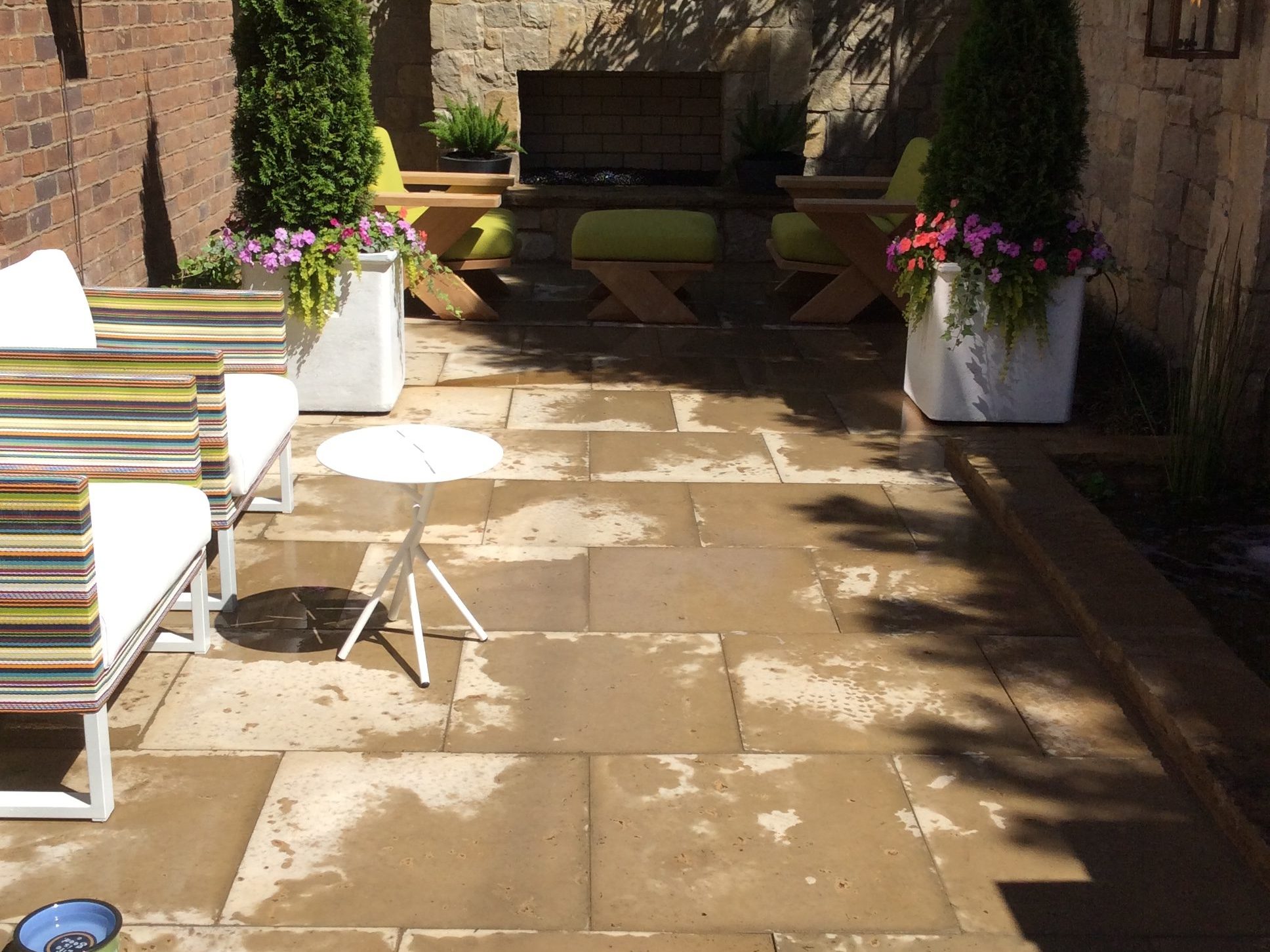 Outdoor patio with stone tiles, a small round table, striped chairs, planters with flowers, and a brick fireplace at the back.
