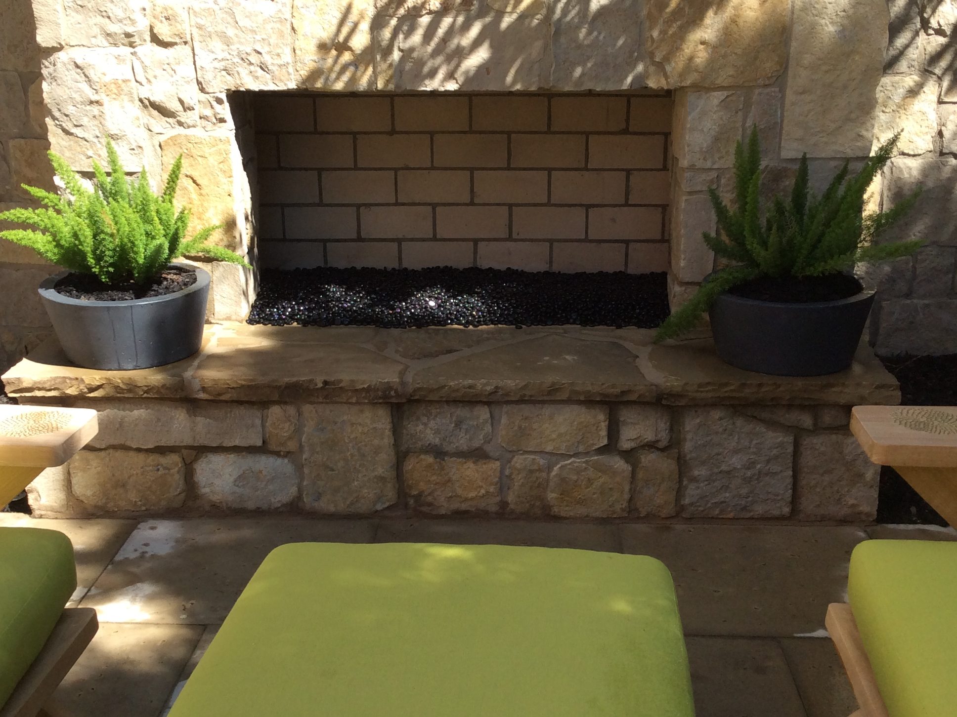 Outdoor stone fireplace with a brick interior, flanked by two potted ferns. Lime green cushioned ottomans are in the foreground.