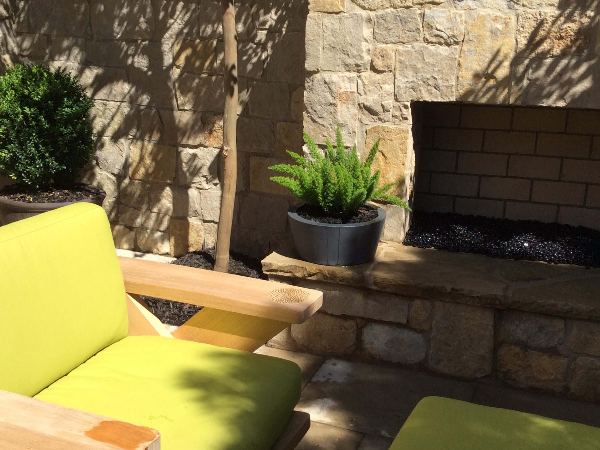 Stone outdoor fireplace with a small potted plant on the ledge. Bright green patio seating is in the foreground, surrounded by sunlit stone walls and a tree.