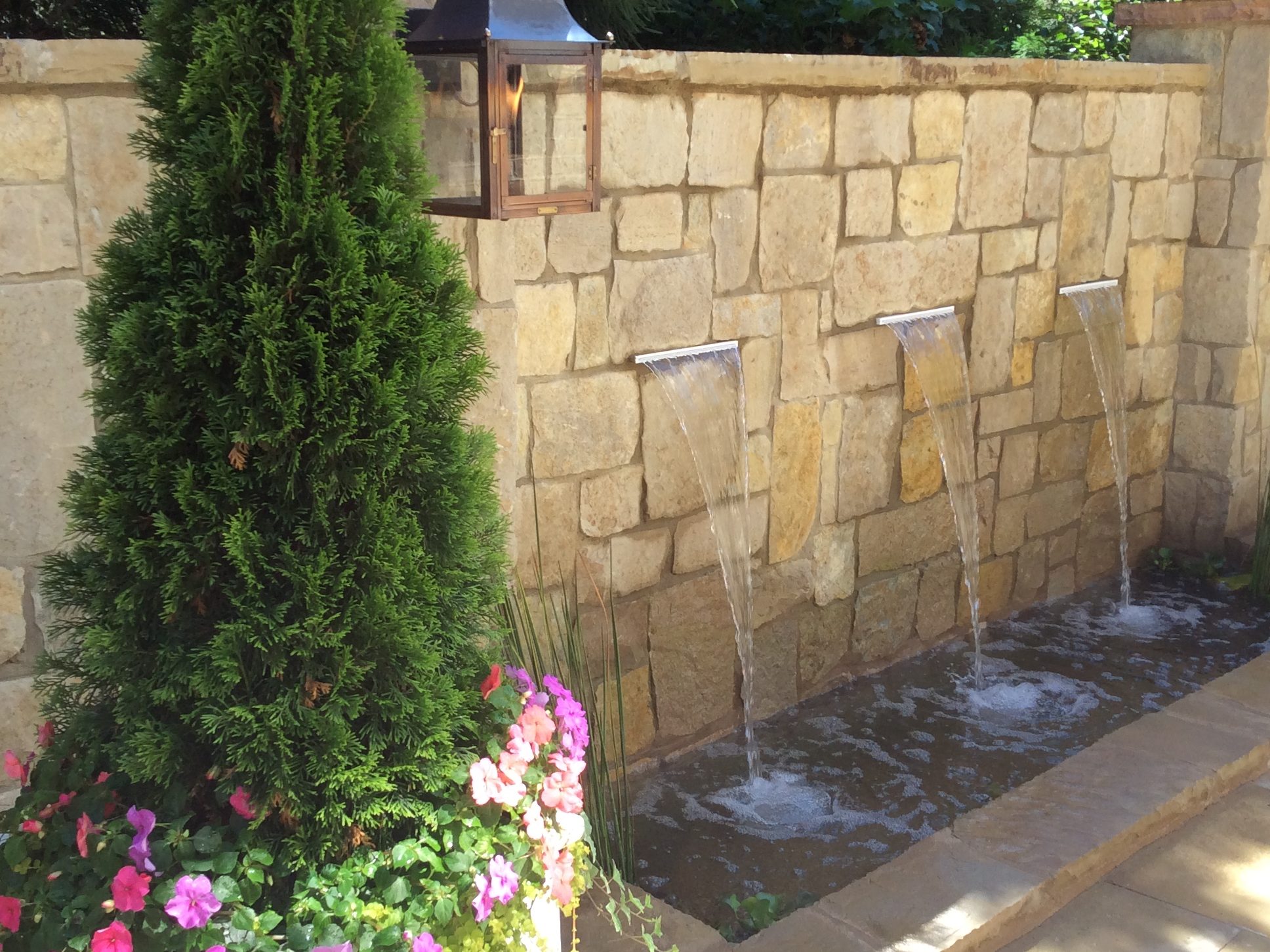A stone water feature with three spouts flows into a shallow pool. A potted plant with pink flowers and a lantern are nearby.
