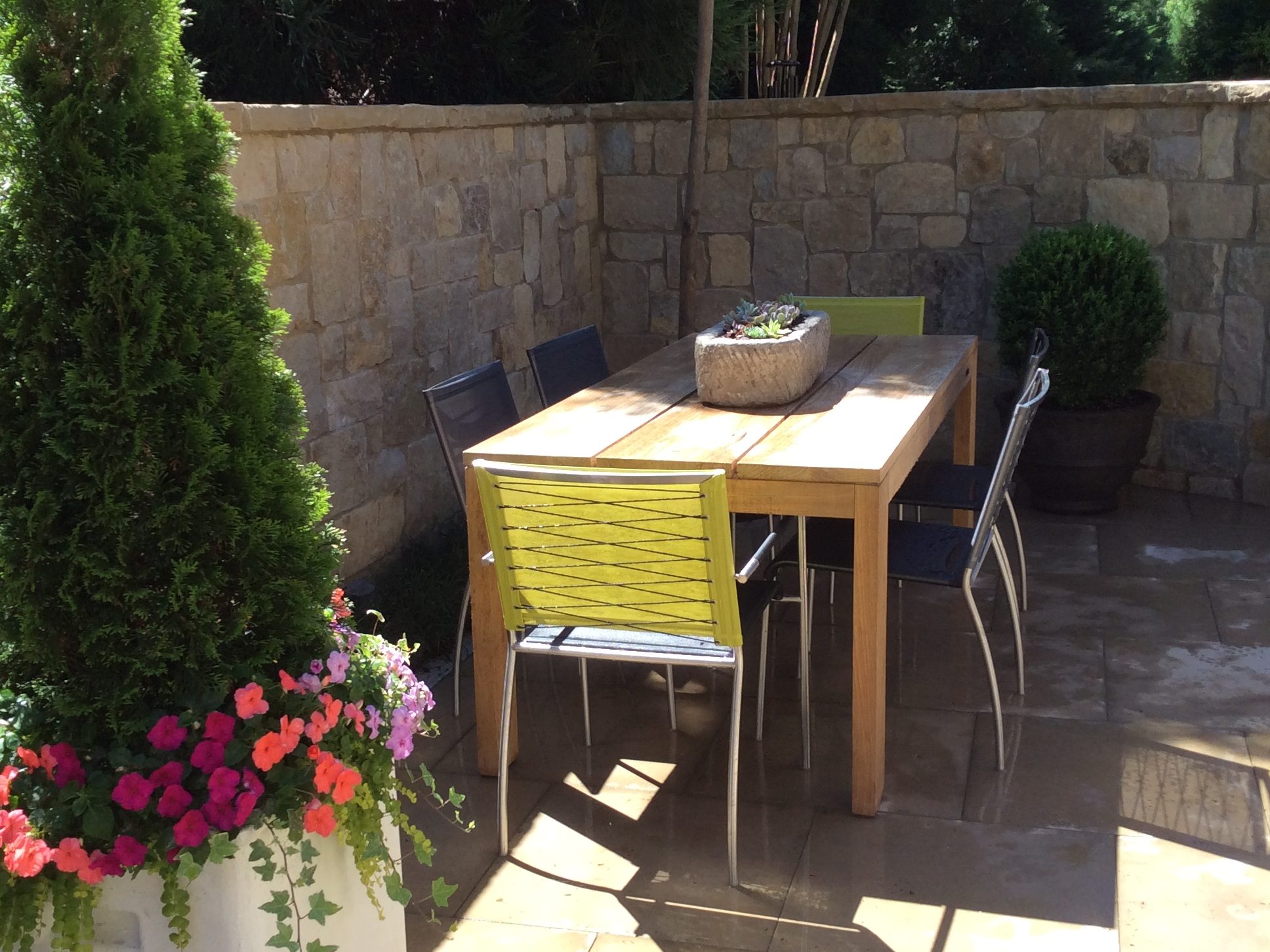 A wooden outdoor dining table with six chairs, surrounded by stone walls. A planter with colorful flowers is in the foreground, and tall greenery is in the background.