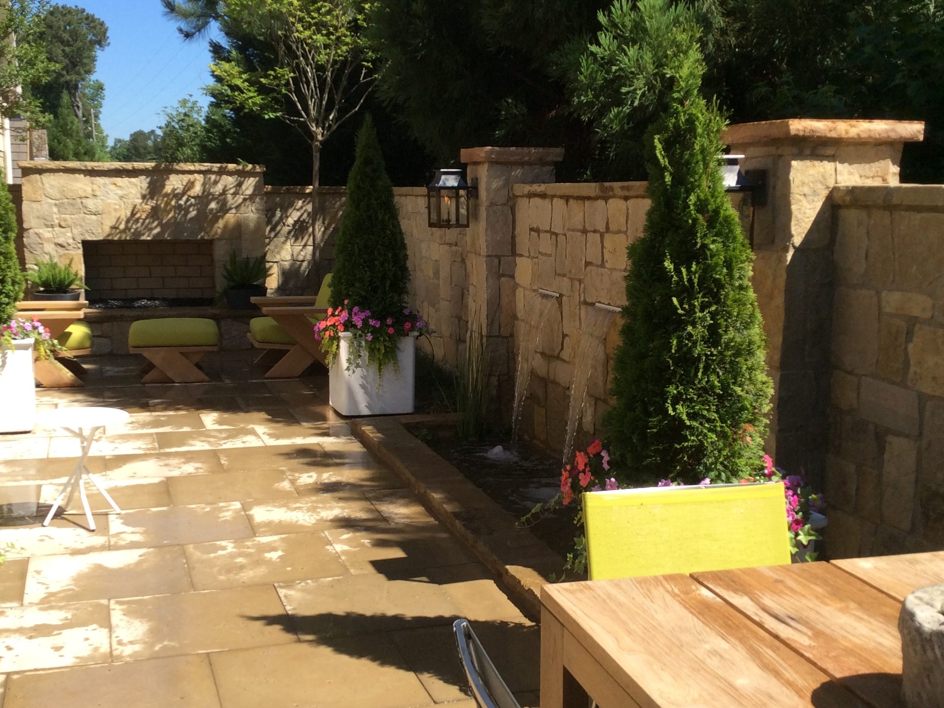 Sunlit patio with stone flooring and walls, featuring potted plants, cushioned benches, and a wooden table.