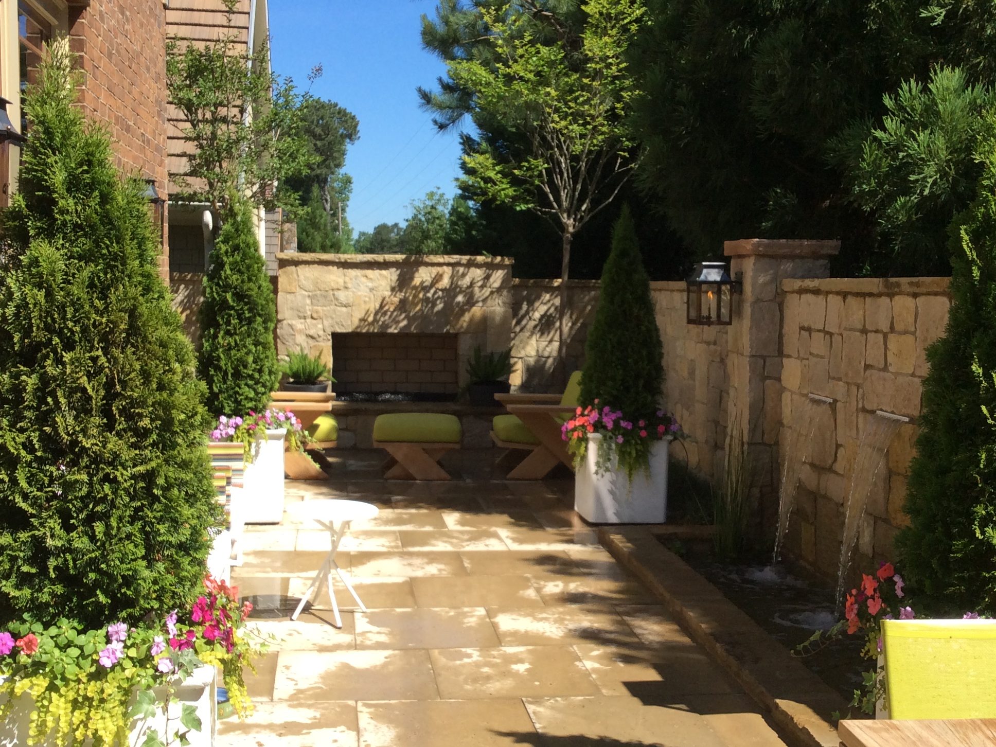 A sunny patio with stone flooring, surrounded by trees and plants in planters, and features benches with green cushions by a stone wall.