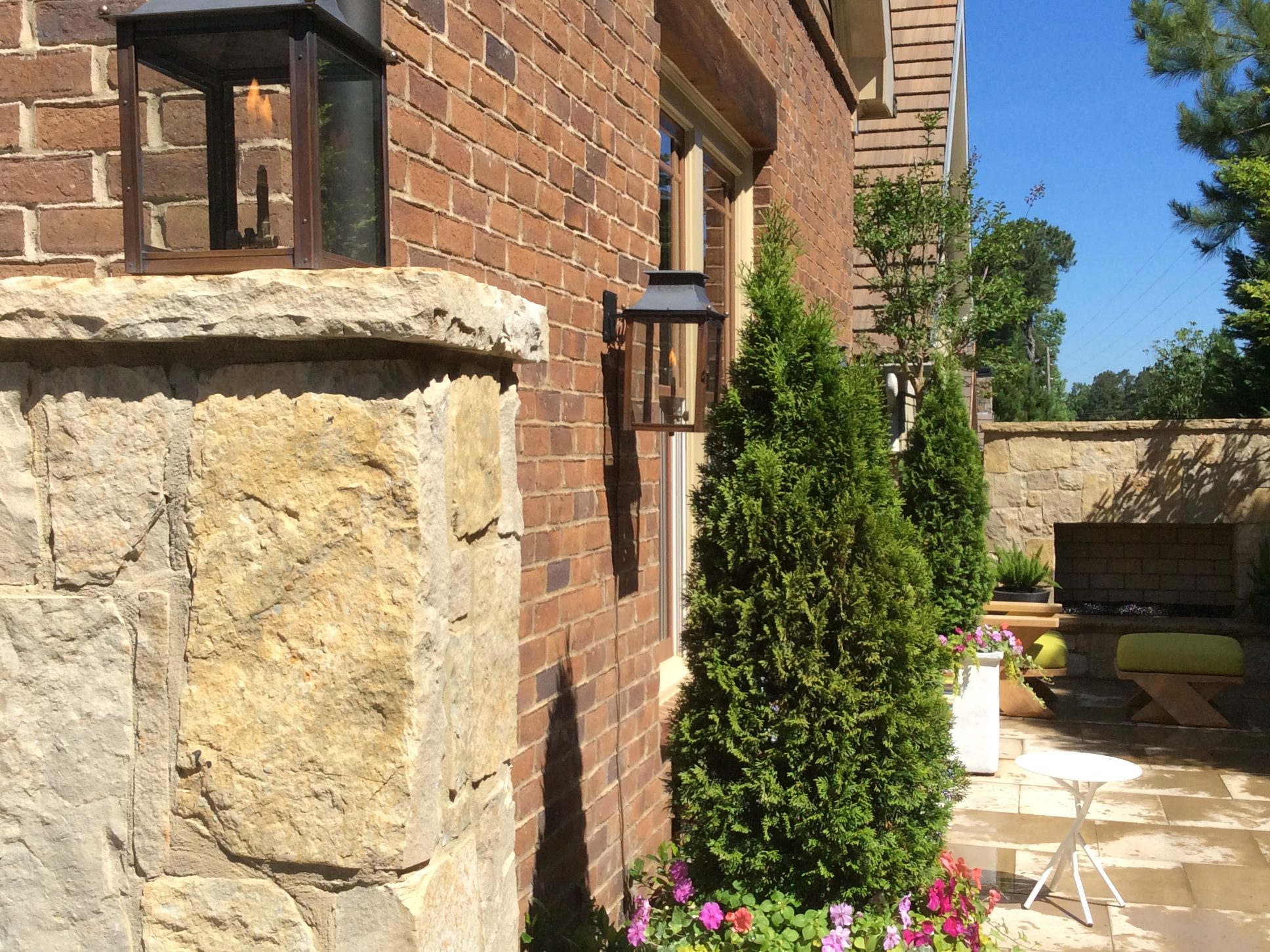 Brick and stone patio with tall potted evergreens and colorful flowers. Two wall-mounted lamps and a lantern visible. Clear blue sky in the background.