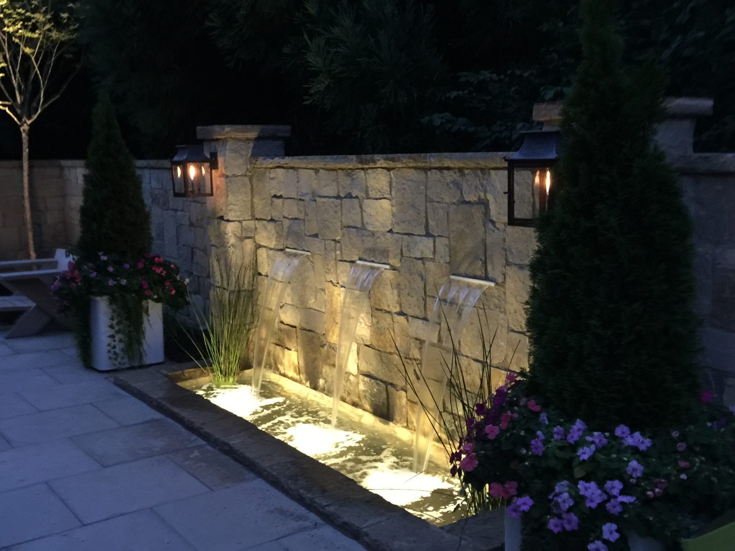 A stone wall features three cascading water spouts illuminated at night, surrounded by planters with evergreens and colorful flowers.
