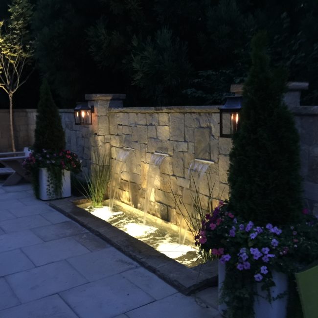 A stone wall features three cascading water spouts illuminated at night, surrounded by planters with evergreens and colorful flowers. A stone wall features three cascading water spouts illuminated at night, surrounded by planters with evergreens and colorful flowers.