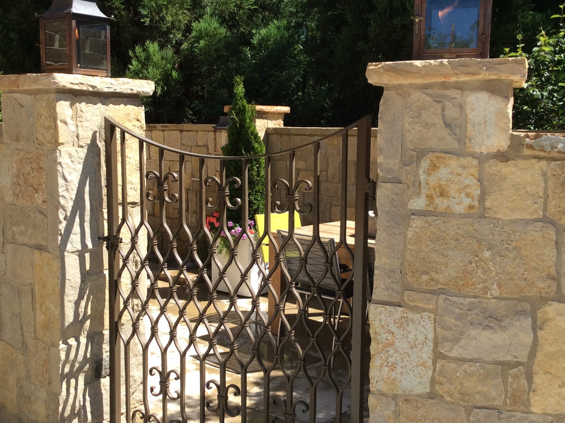 Wrought iron gate with ornate design set between stone pillars, leading to a patio with outdoor furniture and plants. Lanterns are mounted on the pillars.