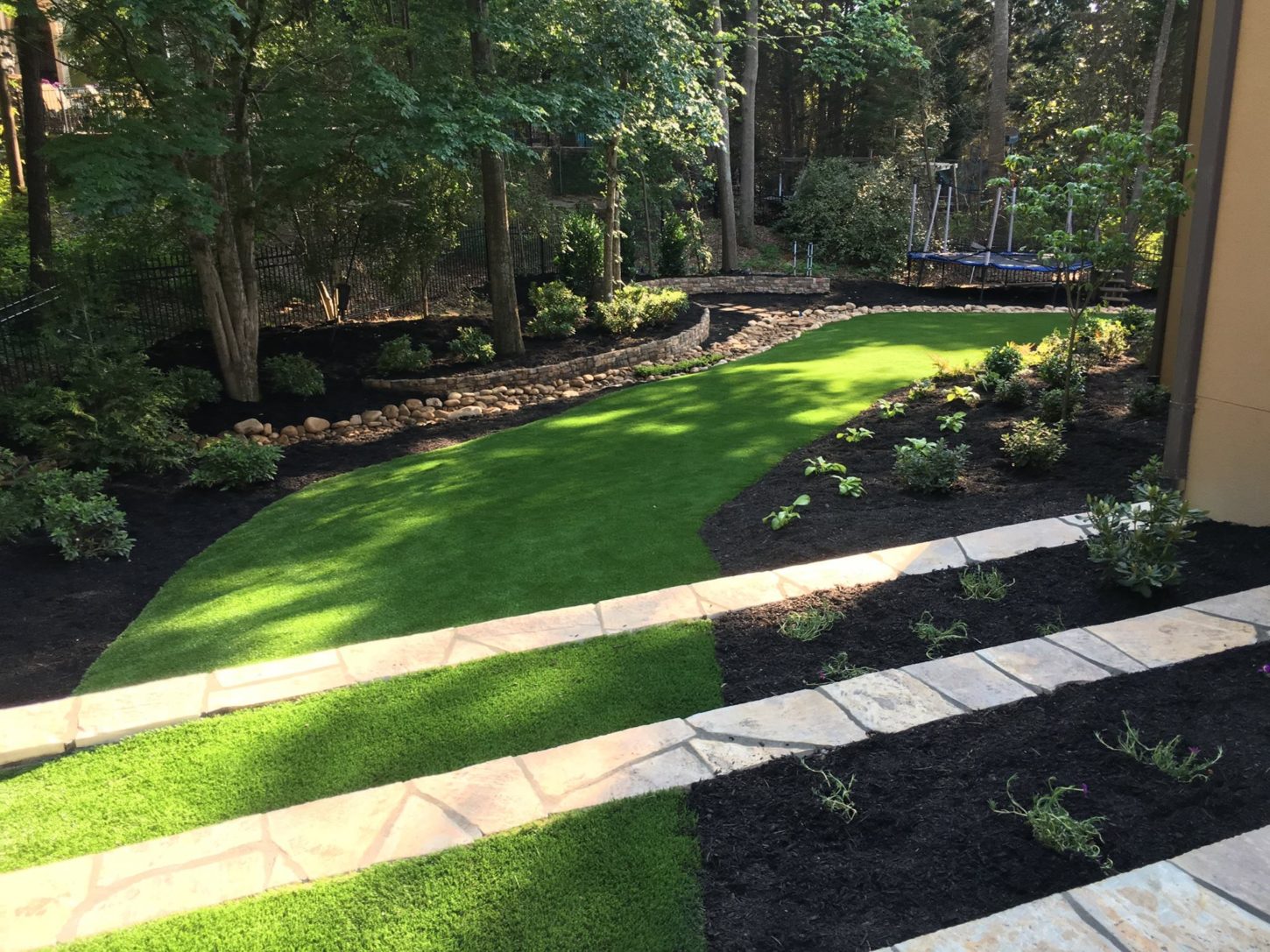 The backyard is a neatly landscaped oasis, complete with a lush green lawn bordered by mulch and vibrant plants. A charming stone pathway meanders through the space, guiding you toward a tranquil wooded area.