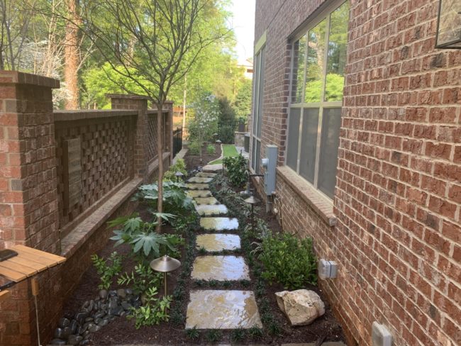 Side yard garden with stepping stones, small plants, and a tree next to a brick house and fence. Side yard garden with stepping stones, small plants, and a tree next to a brick house and fence.