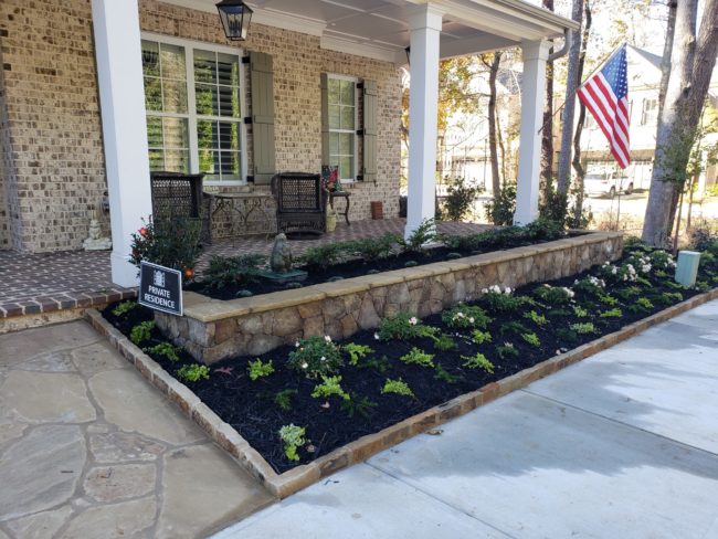 Front yard featuring a raised flower bed and beautiful landscaping, with plants framing the entrance. An American flag flutters above a "Private Residence" sign next to the charming brick house, complete with a porch and inviting chairs.