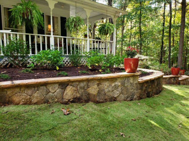 A porch with white railings features hanging plants, overlooking vibrant landscaping that includes a stone retaining wall, red planters, and lush green foliage.