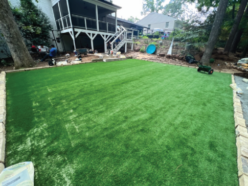 The backyard sanctuary boasts a freshly laid artificial grass lawn, elegantly bordered by stone. A charming wooden deck and lush trees grace the background, creating a serene escape.