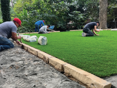 Three people transform the backyard into a serene sanctuary by installing artificial grass, carefully placing bags along one edge.