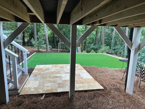View under a deck showcasing stairs, a tiled area, artificial grass, and surrounding trees, creating a tranquil backyard sanctuary.