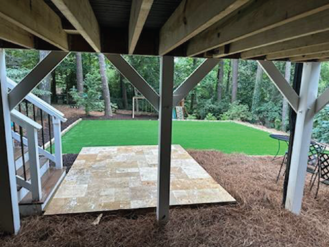 View under a deck showcasing stairs, a tiled area, artificial grass, and surrounding trees, creating a tranquil backyard sanctuary.