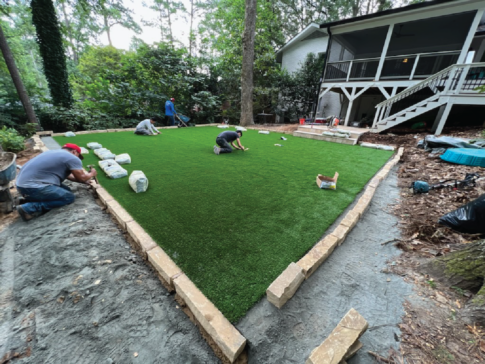Workers are busy transforming a backyard sanctuary by installing artificial grass, bordered by bricks, with trees providing a serene backdrop against the house.