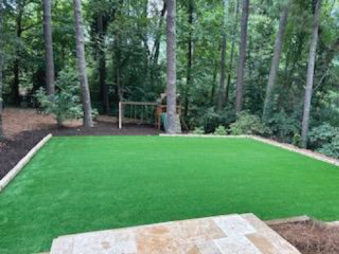 A backyard sanctuary featuring a rectangular patch of artificial grass bordered by trees, with a swing set in the background and stone steps leading down.