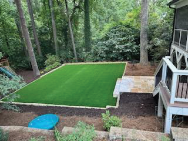 A backyard sanctuary featuring a rectangular artificial grass section is surrounded by lush trees and bushes. A cozy patio nestles beside the house, while a blue kiddie pool adds charm to the corner.