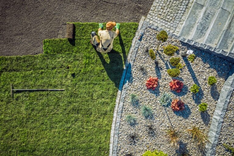 Person laying rolls of sod on a lawn next to a landscaped garden with gravel, small bushes, and stone steps, showcasing expert landscaping design in Atlanta, viewed from above.