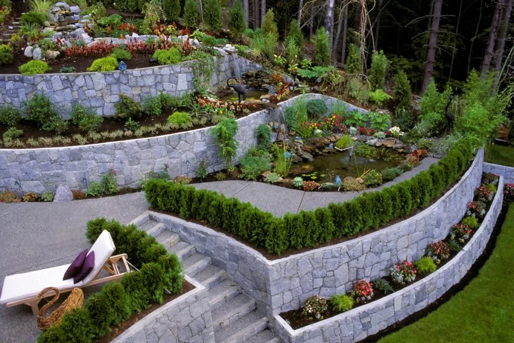 This terraced garden features stone walls resembling those in guides on how to build a retaining wall. It includes a serene pond, diverse plants, and a lounge chair on the patio for ultimate relaxation.