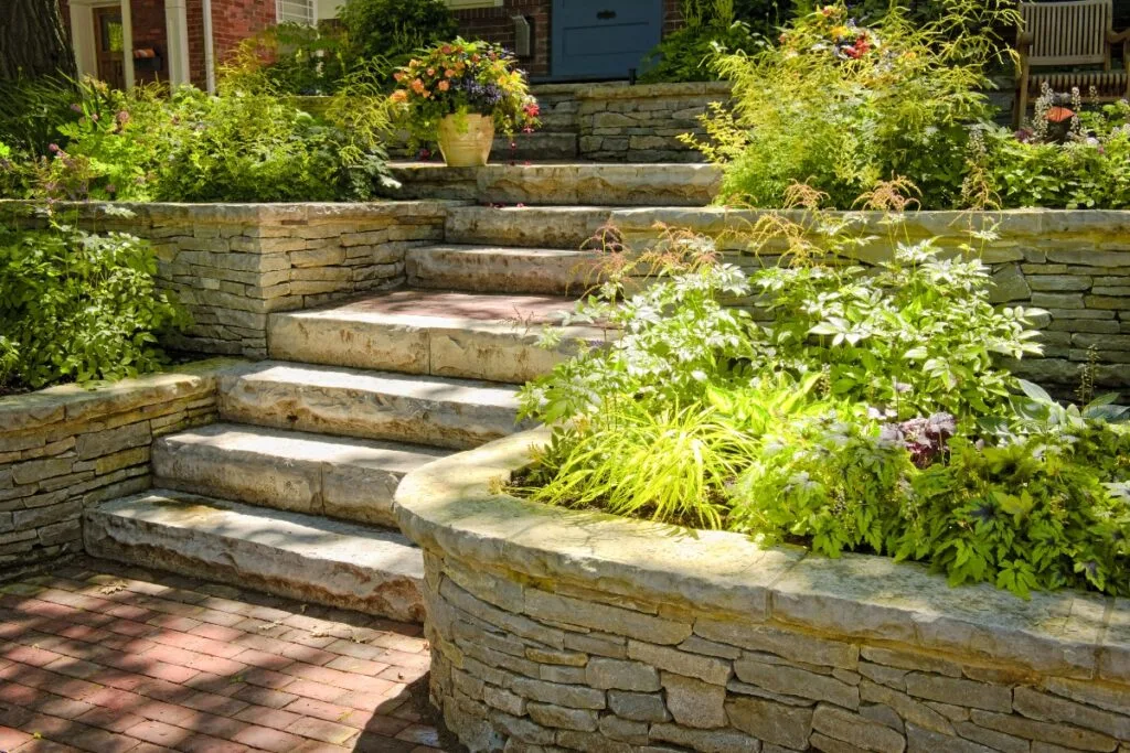 Stone steps with layered garden beds, featuring lush green plants and flowers in a residential outdoor setting, create a picturesque backdrop. Discover how to build a retaining wall to enhance this landscape's structure and charm.