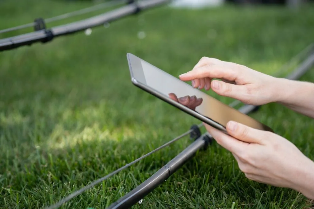 A person relaxes on the grass near a small black tent, using a tablet. They're engrossed in reading about how to design a sprinkler system, blending practicality with nature in their outdoor workspace.