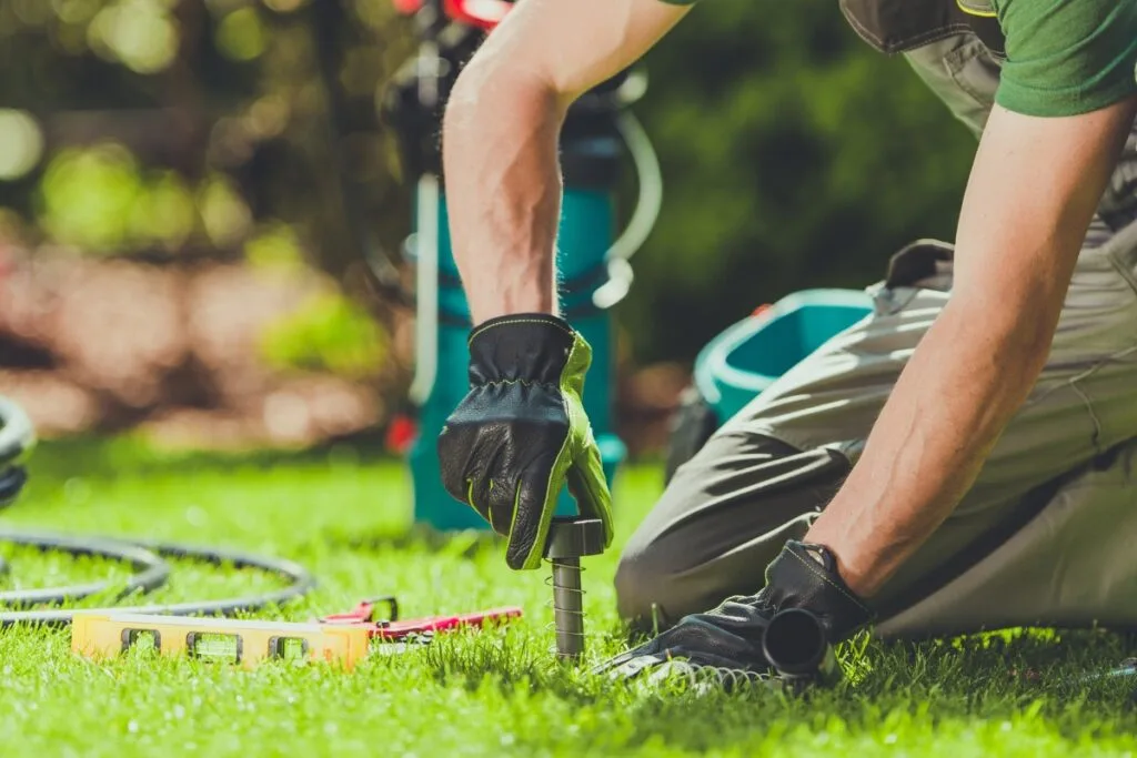 Kneeling on the grass, a person expertly installs a sprinkler system, wearing gloves. Tools and a hose lay beside them—a hands-on moment in learning how to design a sprinkler system.