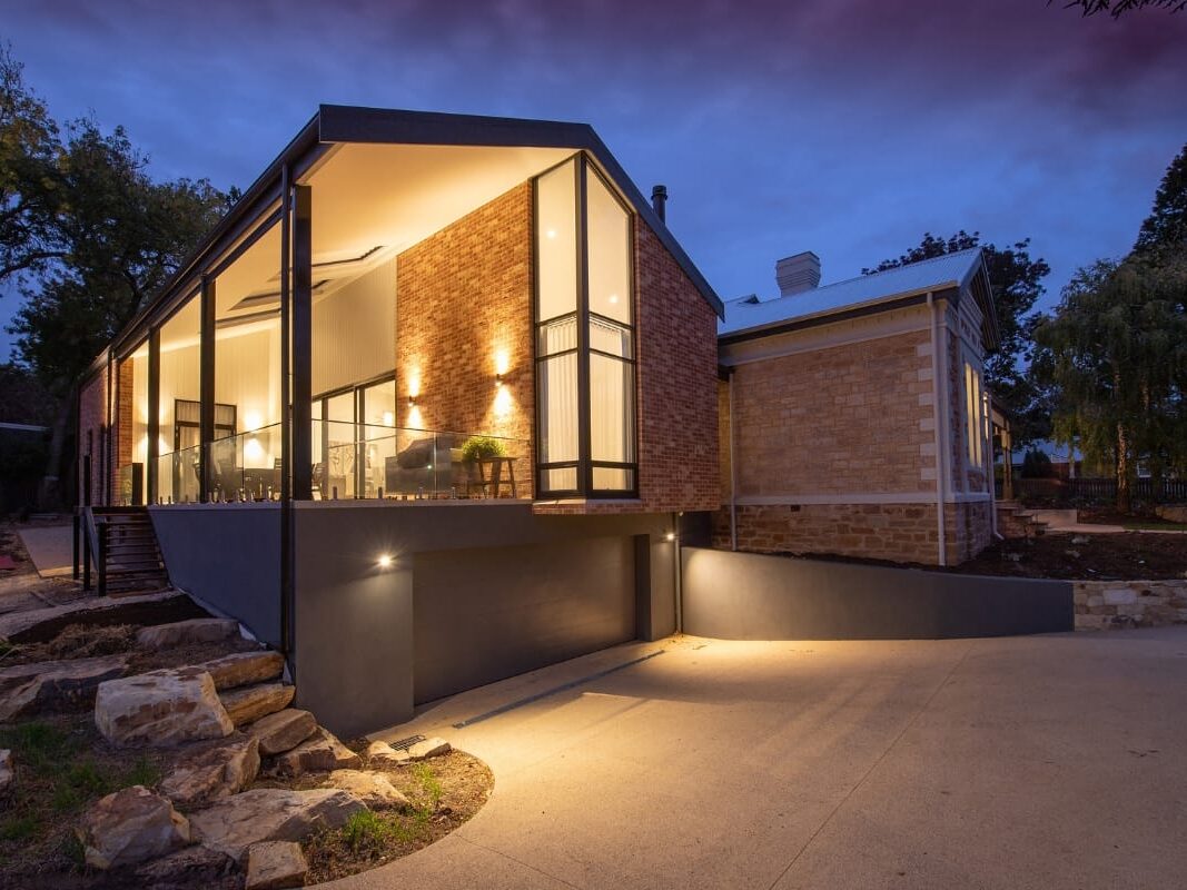Modern house with large glass windows and brick exterior, beautifully illuminated at dusk. Thoughtfully integrated landscape lighting design enhances the stone pathway leading to the entrance, surrounded by trees and elegant stone landscaping.