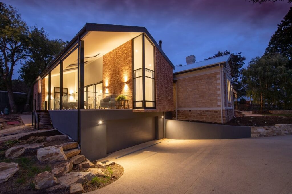 Modern house with large glass windows and brick walls, illuminated at dusk by elegant landscape lighting, featuring a sloped driveway and landscaped stone steps.