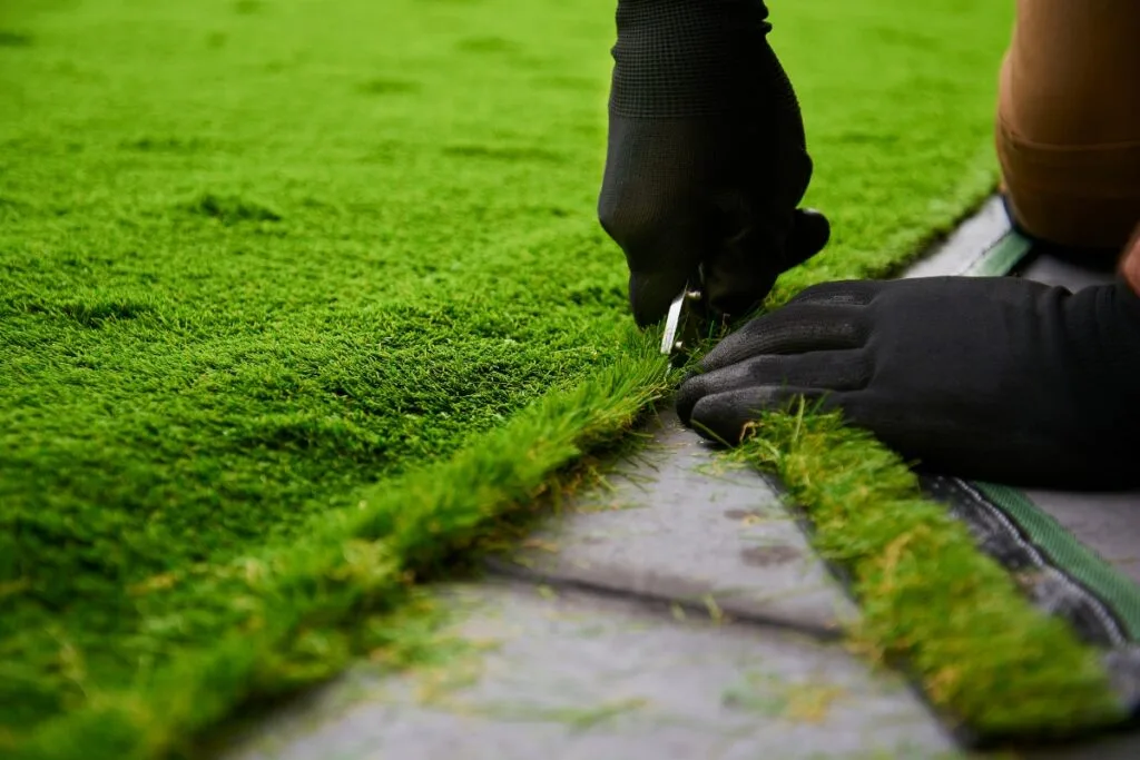 A person wearing gloves is carefully edging the artificial grass with scissors, ensuring every cut is precise and clean.