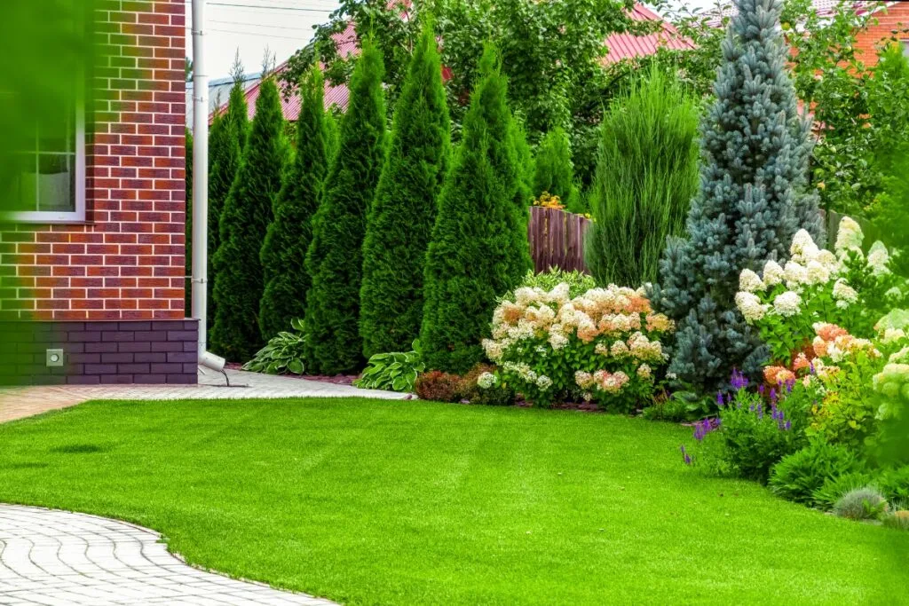 A lush garden with sustainable landscaping in Georgia features a green lawn, blooming hydrangeas, tall evergreens, and a charming brick house on the left.
