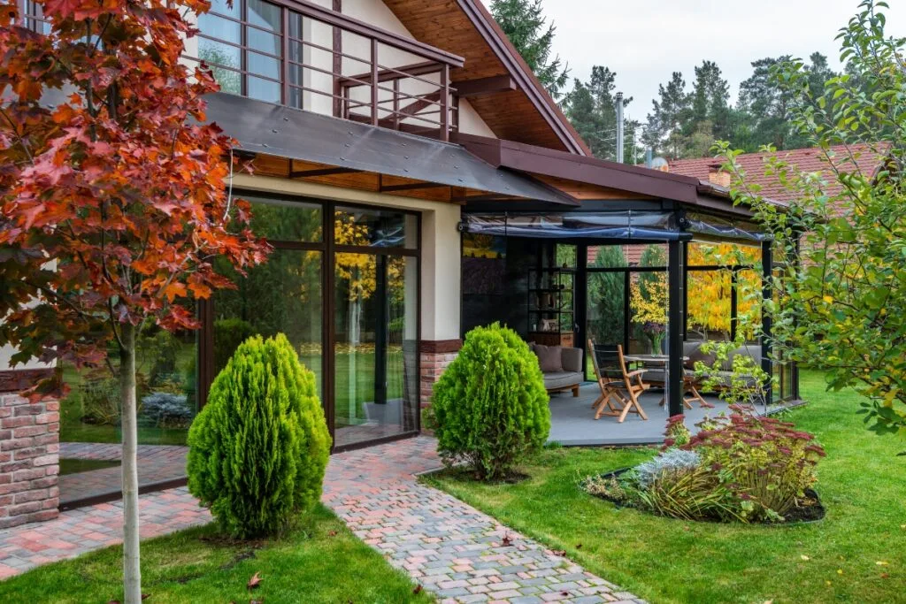 Modern house with large glass windows, surrounded by trees and greenery, showcasing exceptional landscape design in Cherokee County. A patio with a table and chairs is visible, alongside a brick pathway leading through the lush lawn.