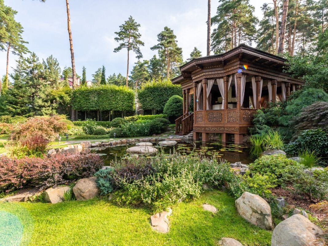 A wooden gazebo by a pond is nestled amidst lush greenery and rocks in this expertly crafted landscape design in Cherokee County. Tall trees form the backdrop under a clear sky, creating an enchanting garden retreat.