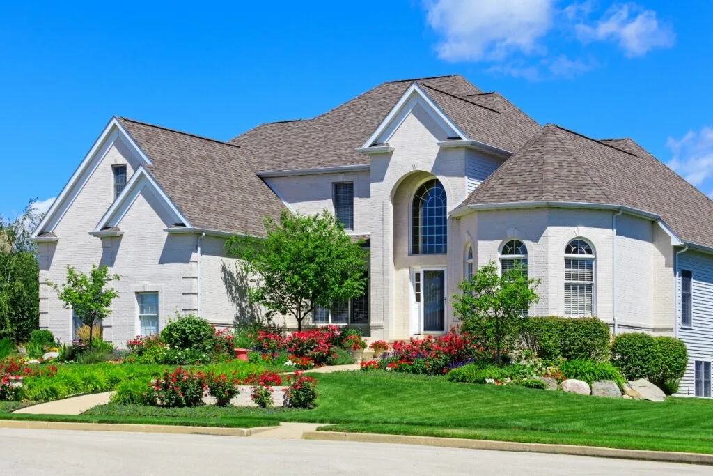 Spacious two-story white brick house with a large arched window, landscaped front yard featuring vibrant red flowers and neatly trimmed shrubs. Enjoy the clear blue sky overhead, all perfectly showcasing the finest landscape design in Cherokee County.