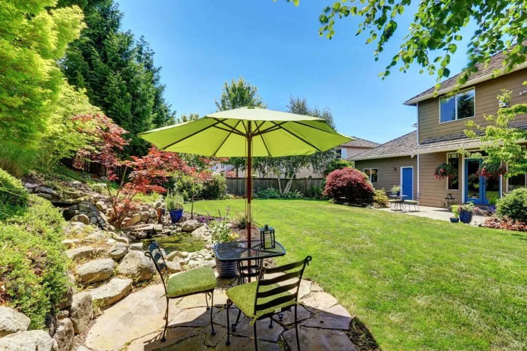 A charming backyard with a green patio umbrella, table, and chairs on a stone surface showcases exceptional landscape design in Cherokee County. It's surrounded by lush plants, trees, and the inviting sight of a two-story house in the background.