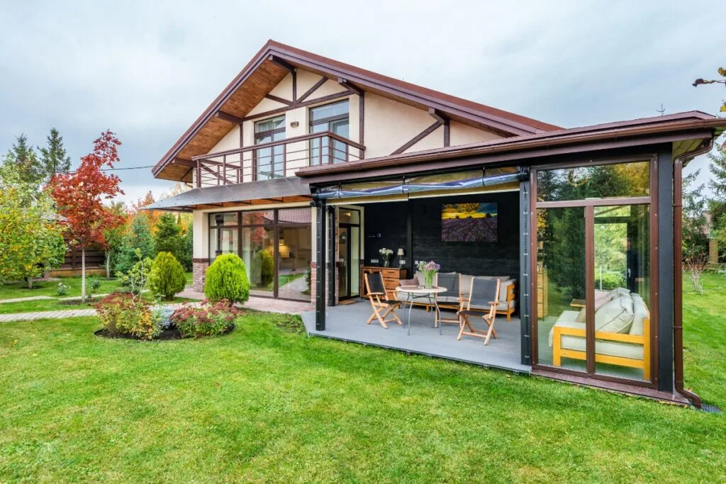 A modern two-story house with a wooden facade, a balcony, and a covered patio showcases exquisite landscape design in Cherokee County. The lawn is neatly kept with small shrubs and a tree, set against a cloudy sky.