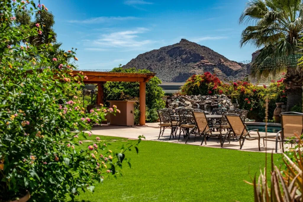 Outdoor patio with a dining table and chairs on a lawn, framed by the lush landscape design in Cherokee County. A stone fountain complements the scene, set against a mountain backdrop under a clear blue sky.