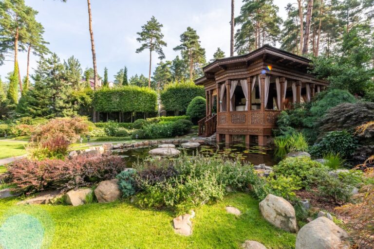 A wooden pavilion with intricate carvings stands beside a pond in a landscaped garden, showcasing custom landscape design that enhances curb appeal in Cherokee County.