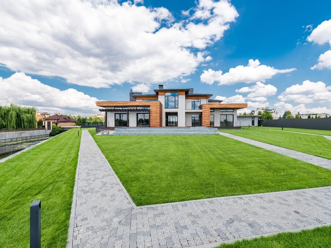 Modern two-story house with large windows and wood accents, surrounded by a luxury landscape design in Georgia, featuring a well-kept lawn and paved walkways under a partly cloudy sky.