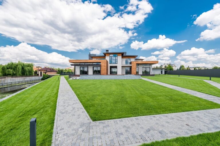 A modern two-story house with large windows and a stone facade sits at the center of a manicured lawn, featuring luxury landscape design and paved walkways under the clear Georgia sky.